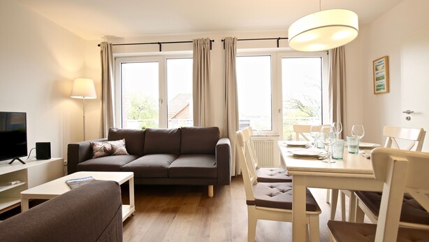 A living room with a sofa, a dining table with chairs, a TV, and large windows with curtains. - Ferienwohnung Leuchtturmblick (Wangerooge)