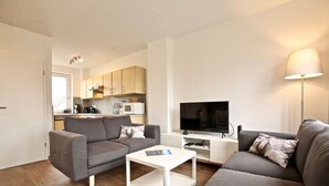 A modern living room with a grey sofa, a white coffee table, and a TV. There's a kitchen in the background with wooden cabinets and a microwave. - Ferienwohnung Leuchtturmblick (Wangerooge)