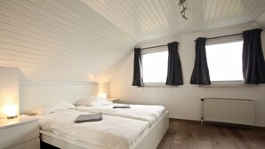 A bedroom with a white bed, nightstands, a window with curtains, and a radiator. - Ferienwohnung Leuchtturmblick (Wangerooge)