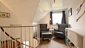 A cozy room with a wooden staircase, two armchairs, a small table, and a bookshelf. - Ferienwohnung Leuchtturmblick (Wangerooge)