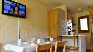 A dining area with a table set for four, a television mounted on the wall, wooden chairs, and a kitchen area with cabinets and a refrigerator. - Grand Cap 4 - Appt 4 pers in Chalet - CALM & NATURE (Tignes)