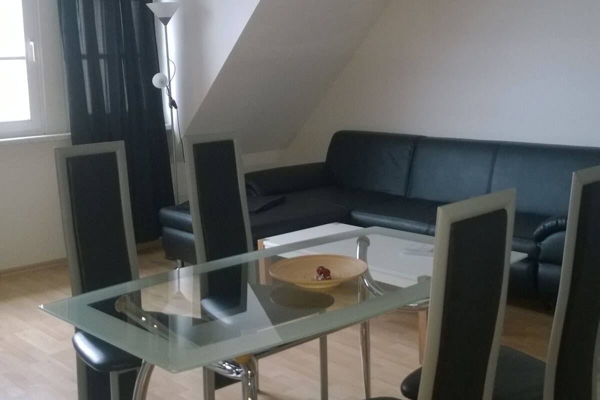 A dining area with a glass table and chairs, a black sofa, and a lamp.