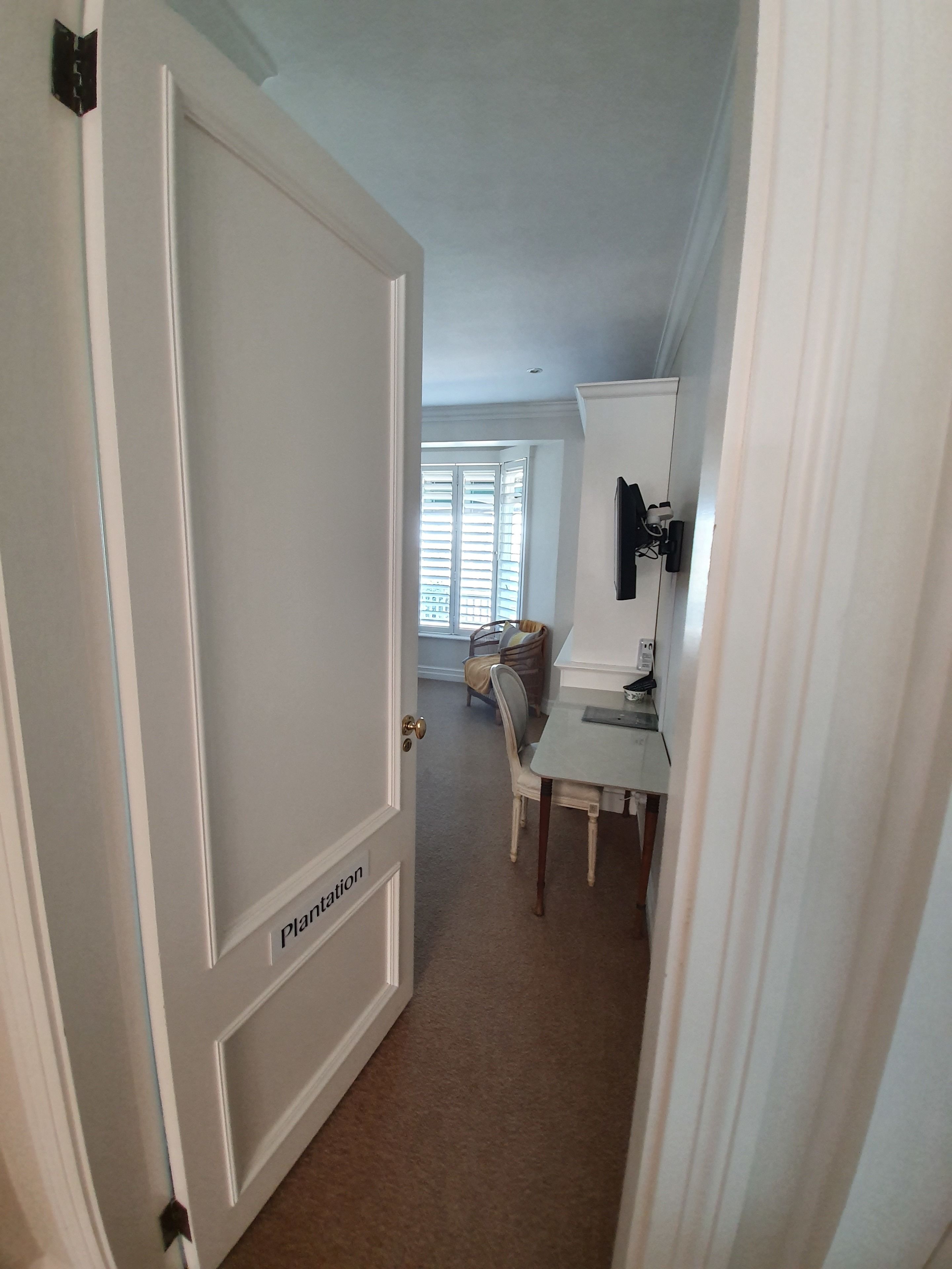 A hallway with a closed door labeled "Plantation," a small table with a chair, a window with blinds, and a mounted TV.
