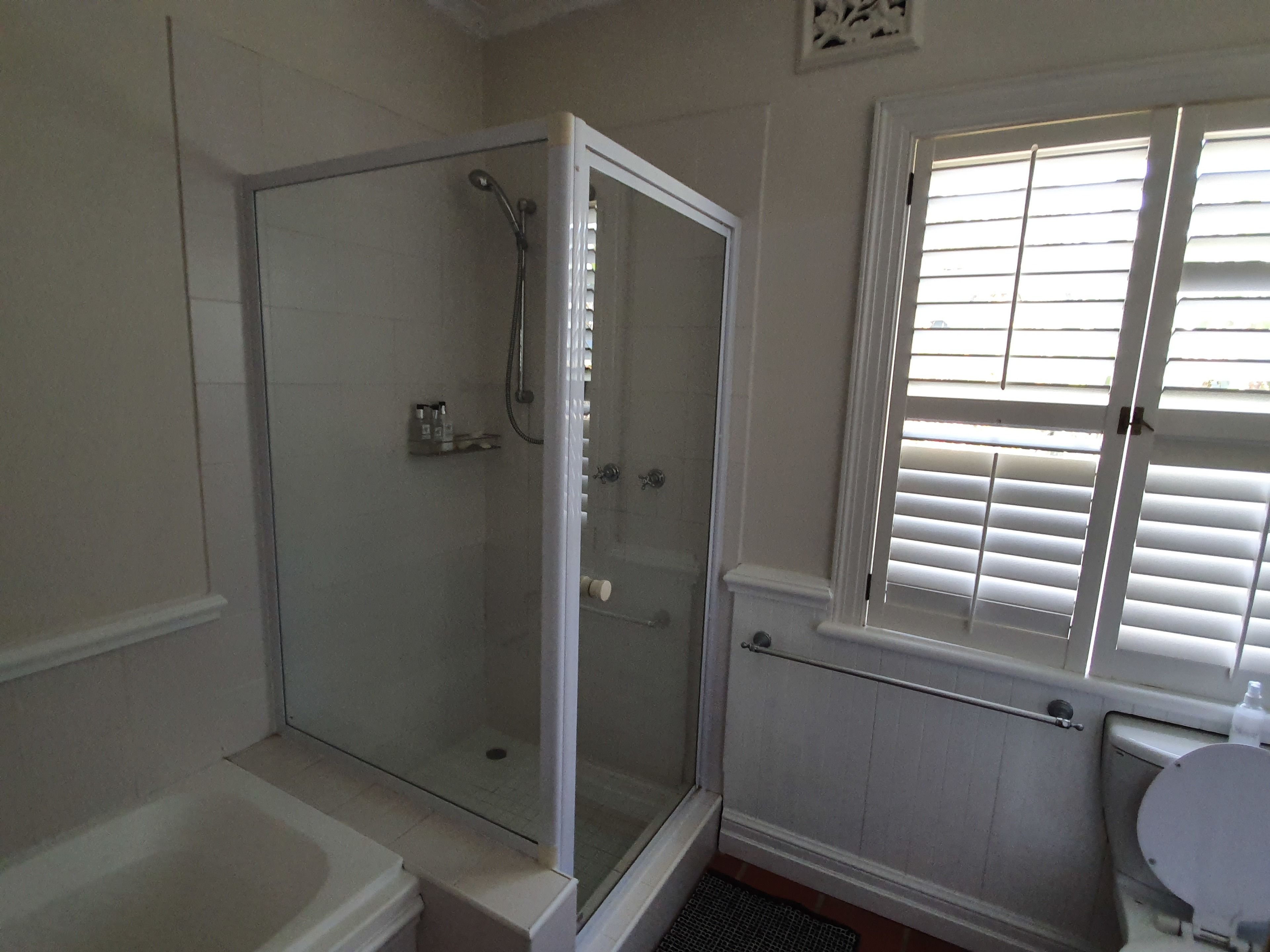 A bathroom with a glass-enclosed shower, a bathtub, a toilet, and a window with blinds.
