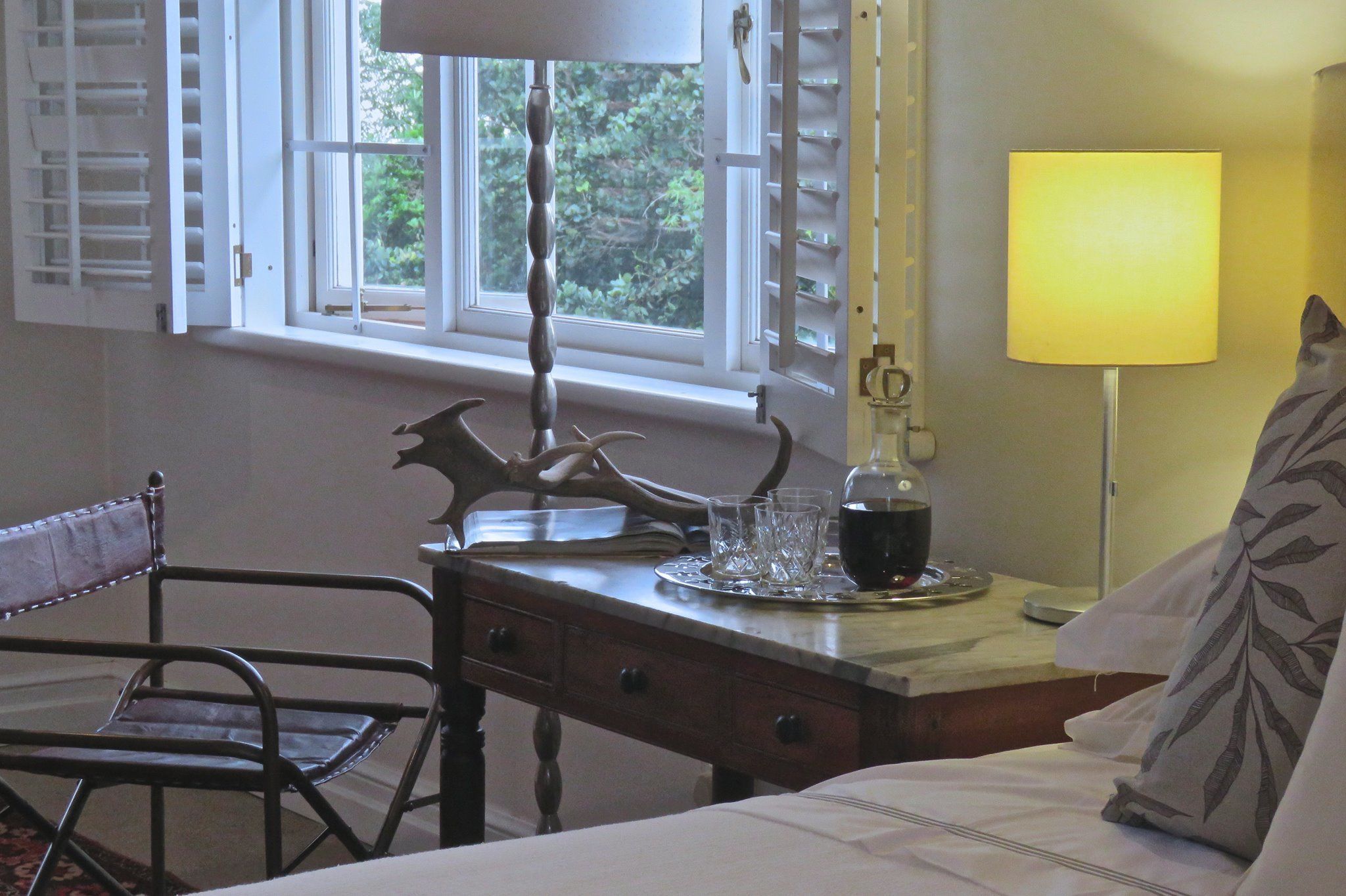 A room with a bed, a wooden desk, a chair, a window with shutters, and a lamp.