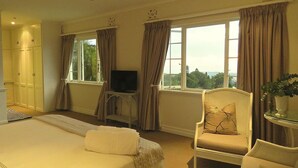 A hotel room with a bed, a TV on a small table, a chair with a cushion, and a window with curtains. - Southey House (Kloof)