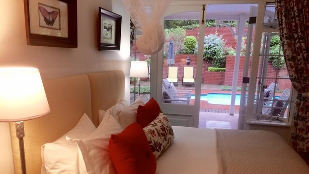 A hotel room with a bed, pillows, a lamp, and a view of a pool and garden. - Southey House (Kloof)