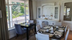 A dining area with a table set for four, a white cabinet with glass doors, and a view of greenery outside. - Southey House (Kloof)