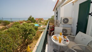 A balcony with a table set for two, overlooking a pool and beach. - Xi Village (Kefalonia)