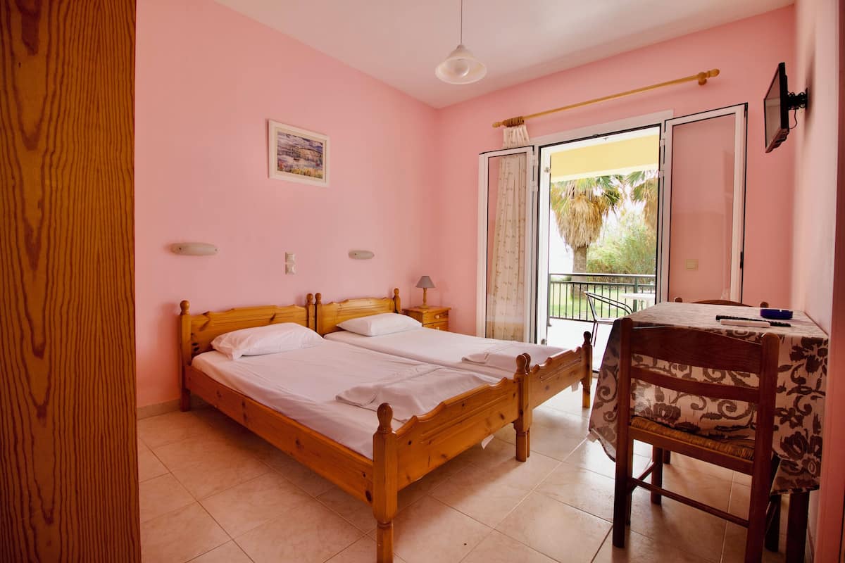 A bedroom with a double bed, a small table with a chair, a window with a view of palm trees, and a door leading to another room.