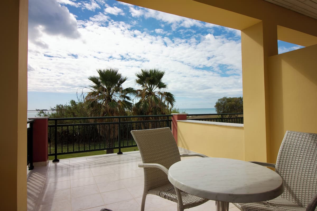 A balcony with wicker chairs and a round table, overlooking a beach with palm trees and a clear sky.