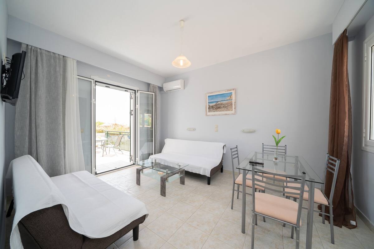 A modern hotel room with a glass dining table, two beds, a TV, and a balcony with a view.