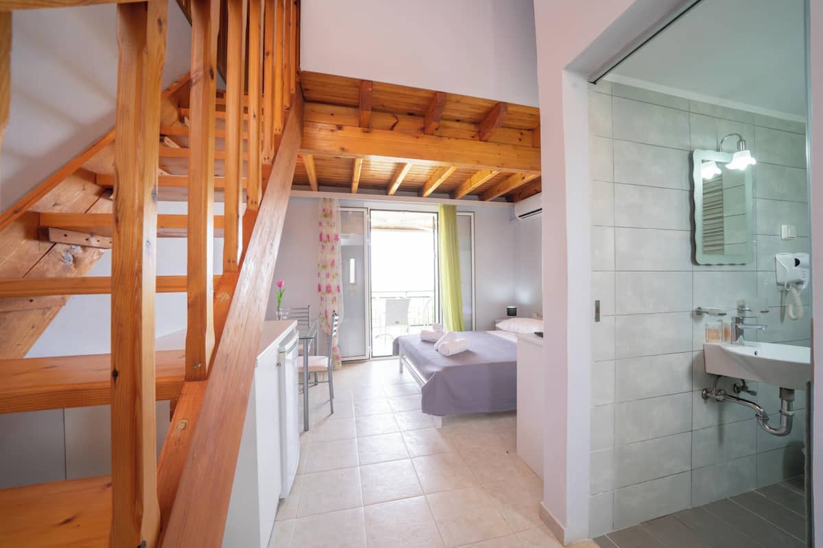 A bedroom with a wooden staircase, a bed, a bathroom with a sink and mirror, and a view of the outside through a window.