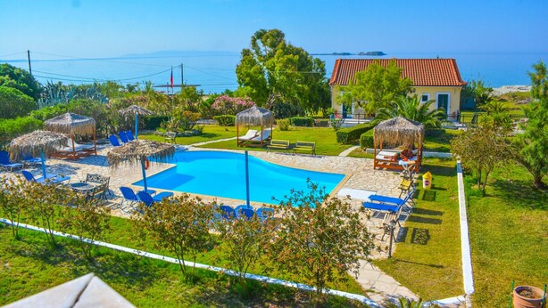 A pool area with lounge chairs, a thatched-roof structure, and a building with a red roof. - Xi Village (Kefalonia)