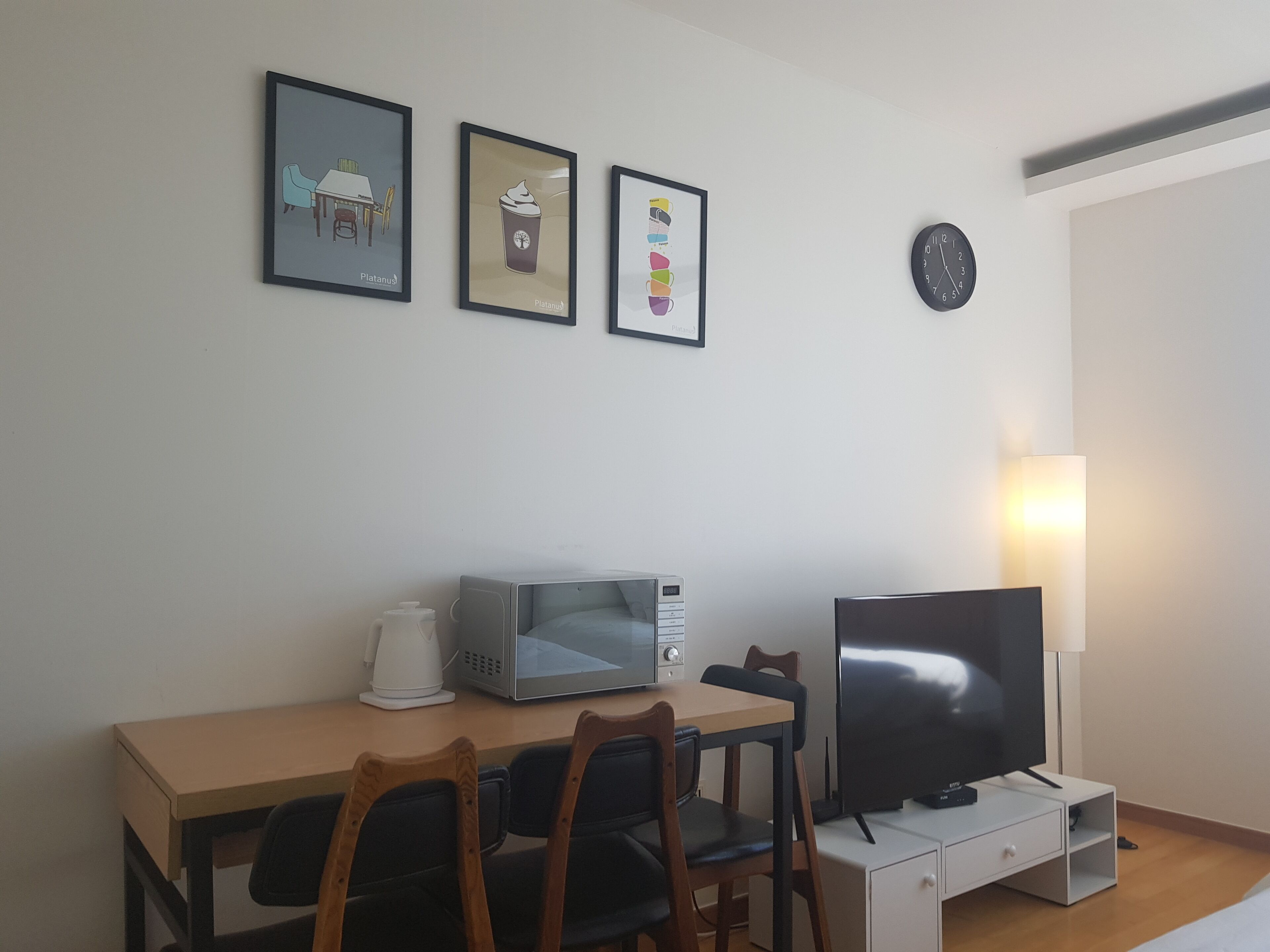 A small, modern room with a wooden desk, a flat-screen TV, a microwave, and a coffee maker. There are three framed pictures on the wall and a clock.