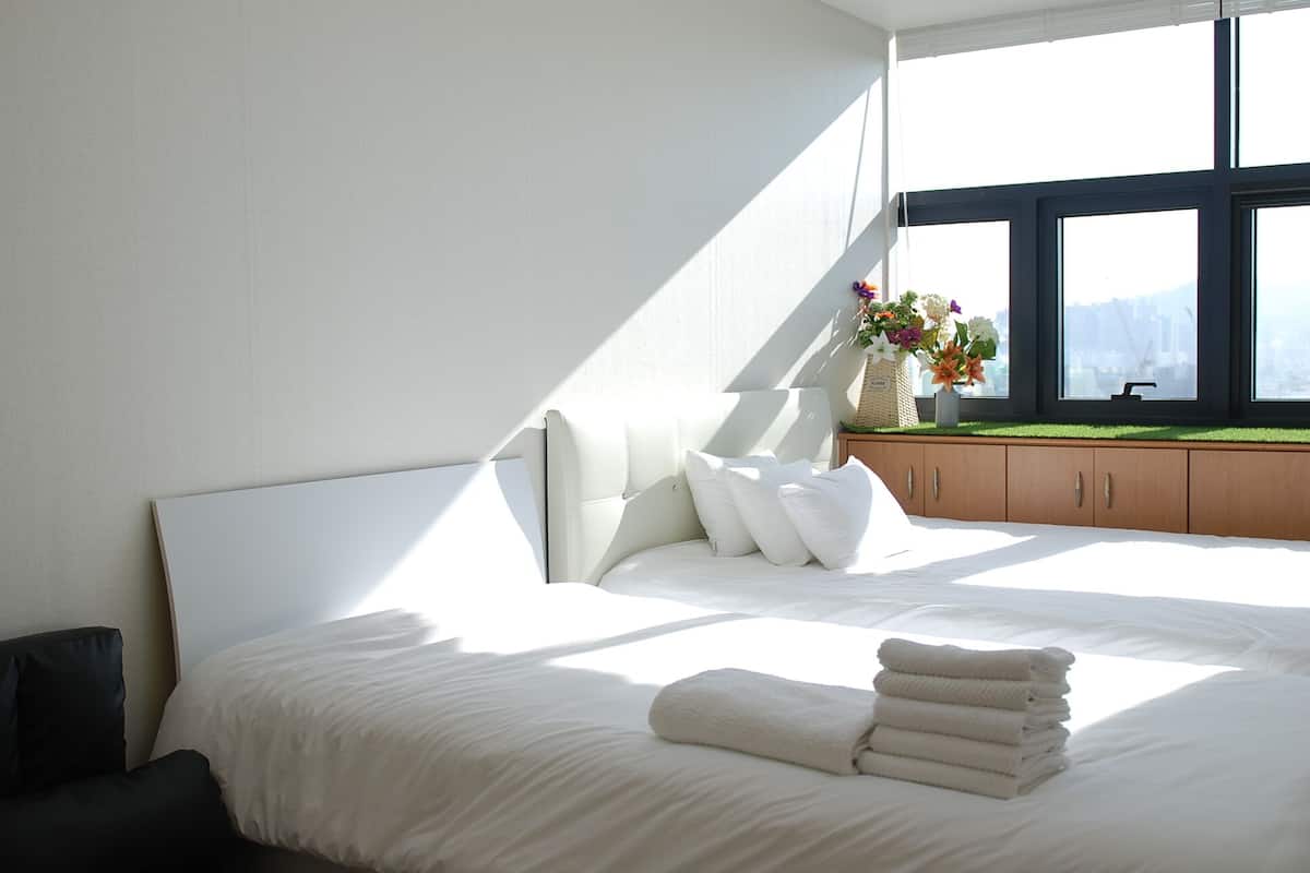 A modern bedroom with a large bed, white linens, a wooden nightstand with a vase of flowers, and a window with a city view.