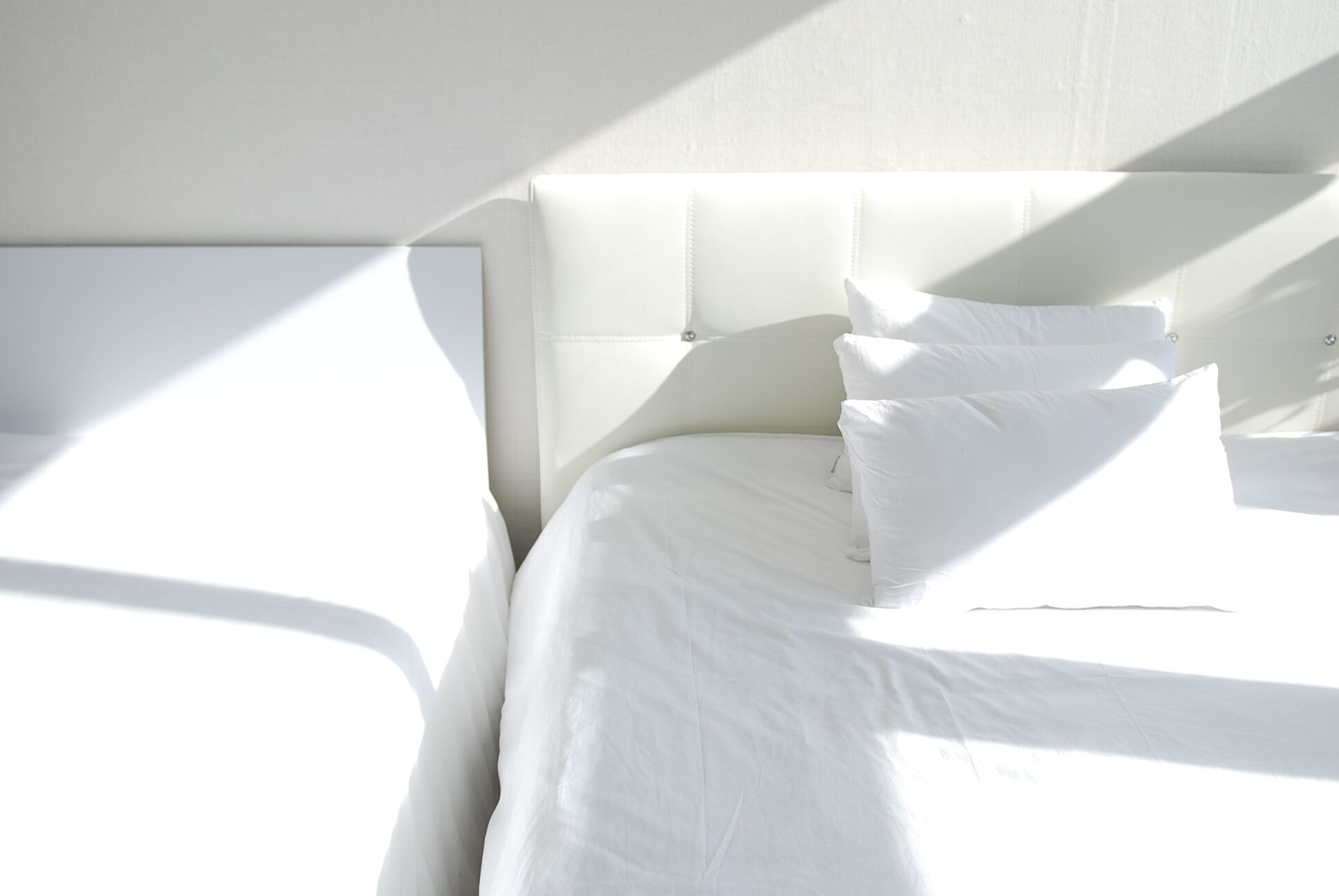 Ein ordentlich gemachtes Bett mit weißen Leinen und Kissen, ein gepolstertes Kopfholz und ein heller, sonnendurchfluteter Raum.