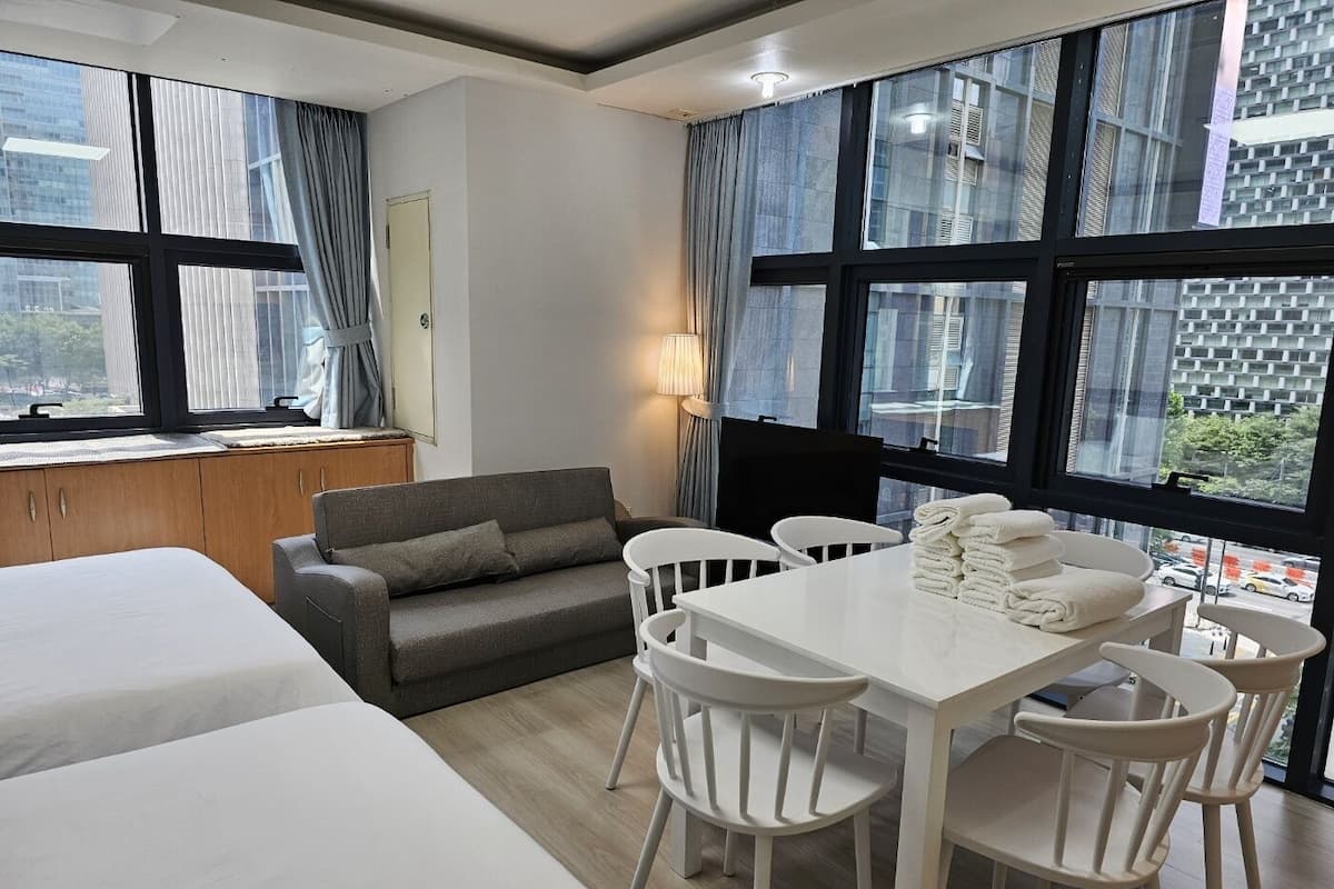 A hotel room with a bed, a sofa, a dining table with chairs, and large windows offering a view of the city.