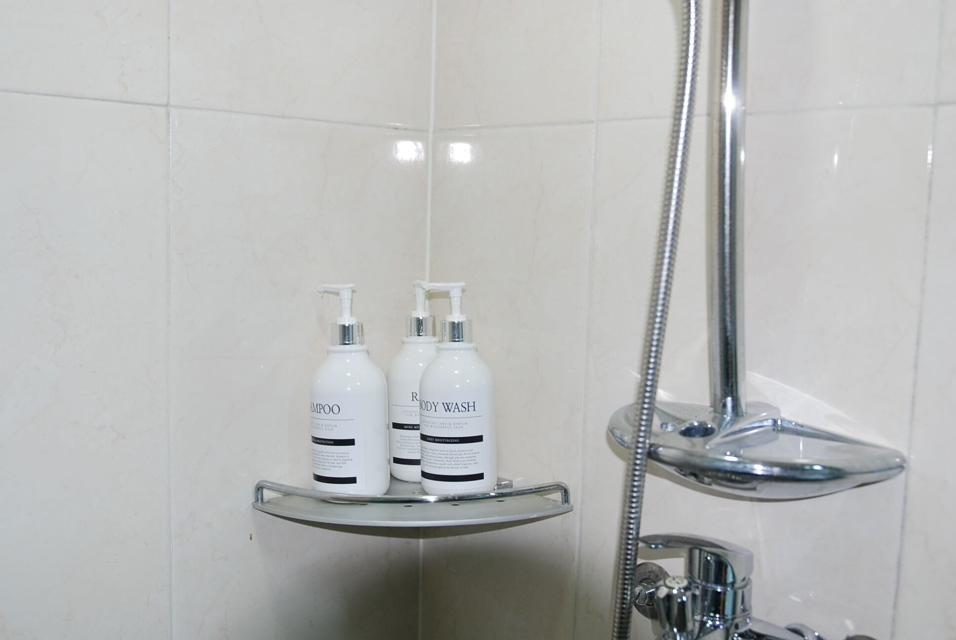 A bathroom with white tiles, a glass shower shelf holding two bottles of shampoo and body wash, and a chrome showerhead.