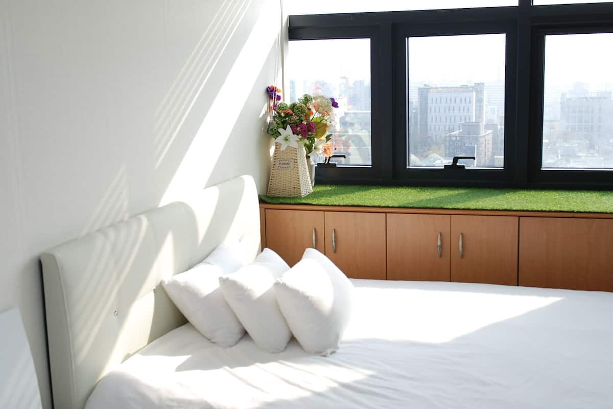 A bedroom with a bed, white pillows, a window with a view of buildings, and a small window seat with a basket and flowers.
