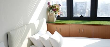 Un dormitorio con cama, almohadas blancas, un ventanal con vistas a edificios y un pequeño rincón con cojín, cesta y flores.