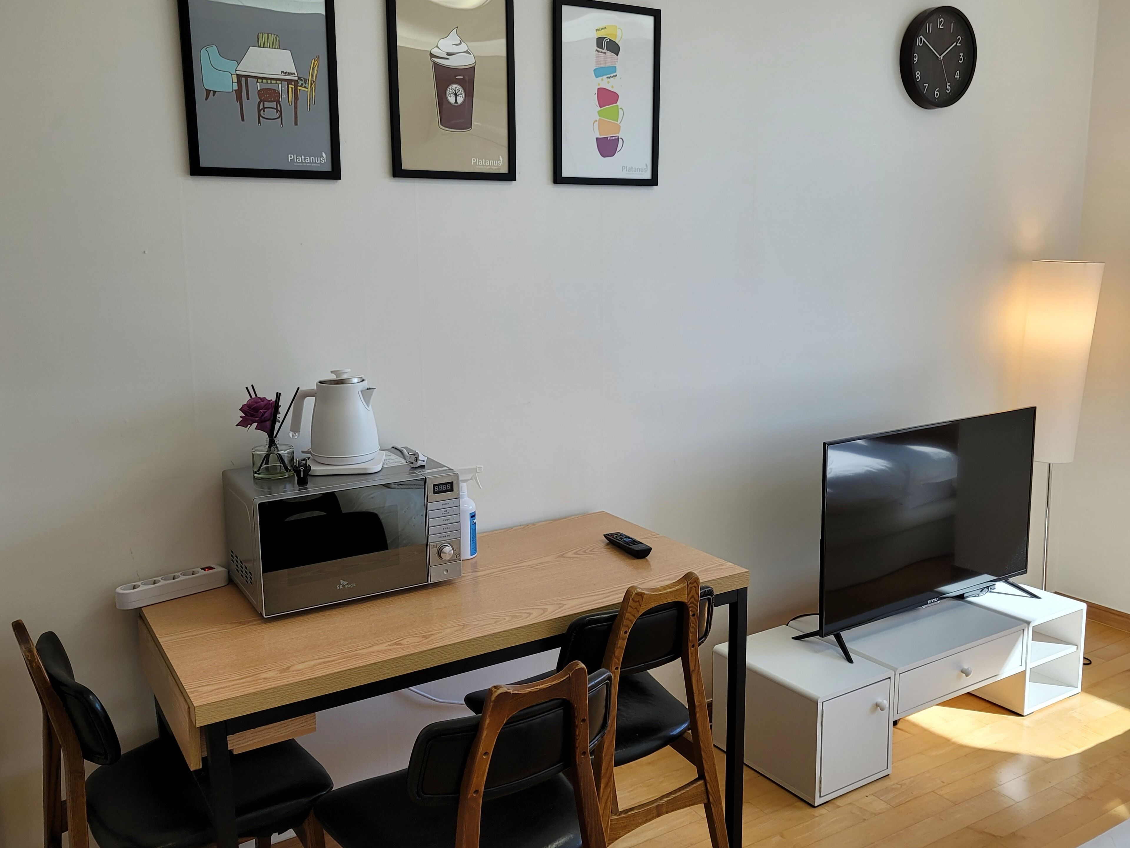 Een kleine, verzorgde kamer met een houten tafel, een magnetron, een televisie en een klok aan de muur.