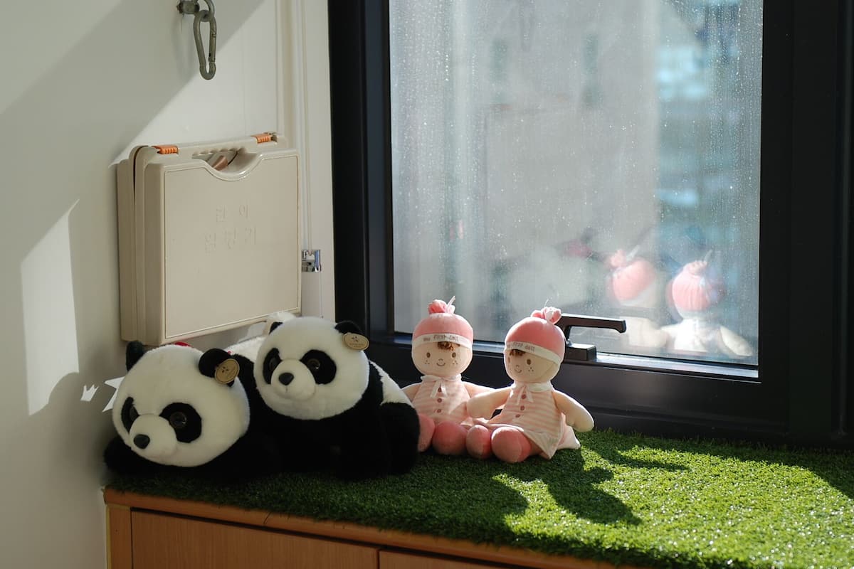 A window with a view of a building and a window sill with two panda plush toys and two dolls.