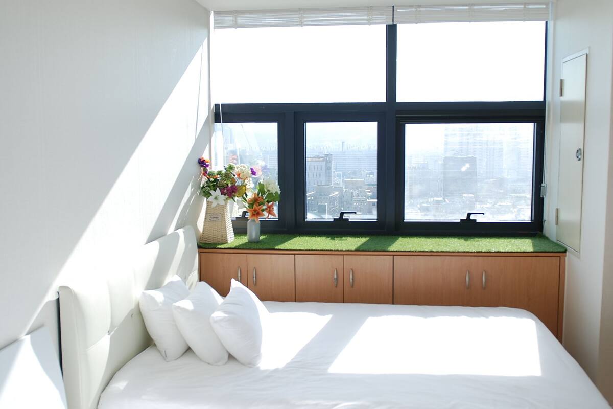 A bedroom with a large window, a bed with white bedding, a wooden cabinet, and a vase with flowers on the windowsill.