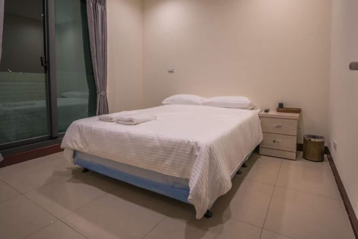 A neatly made bed with a white cover, a bedside table with a lamp, and a trash can.