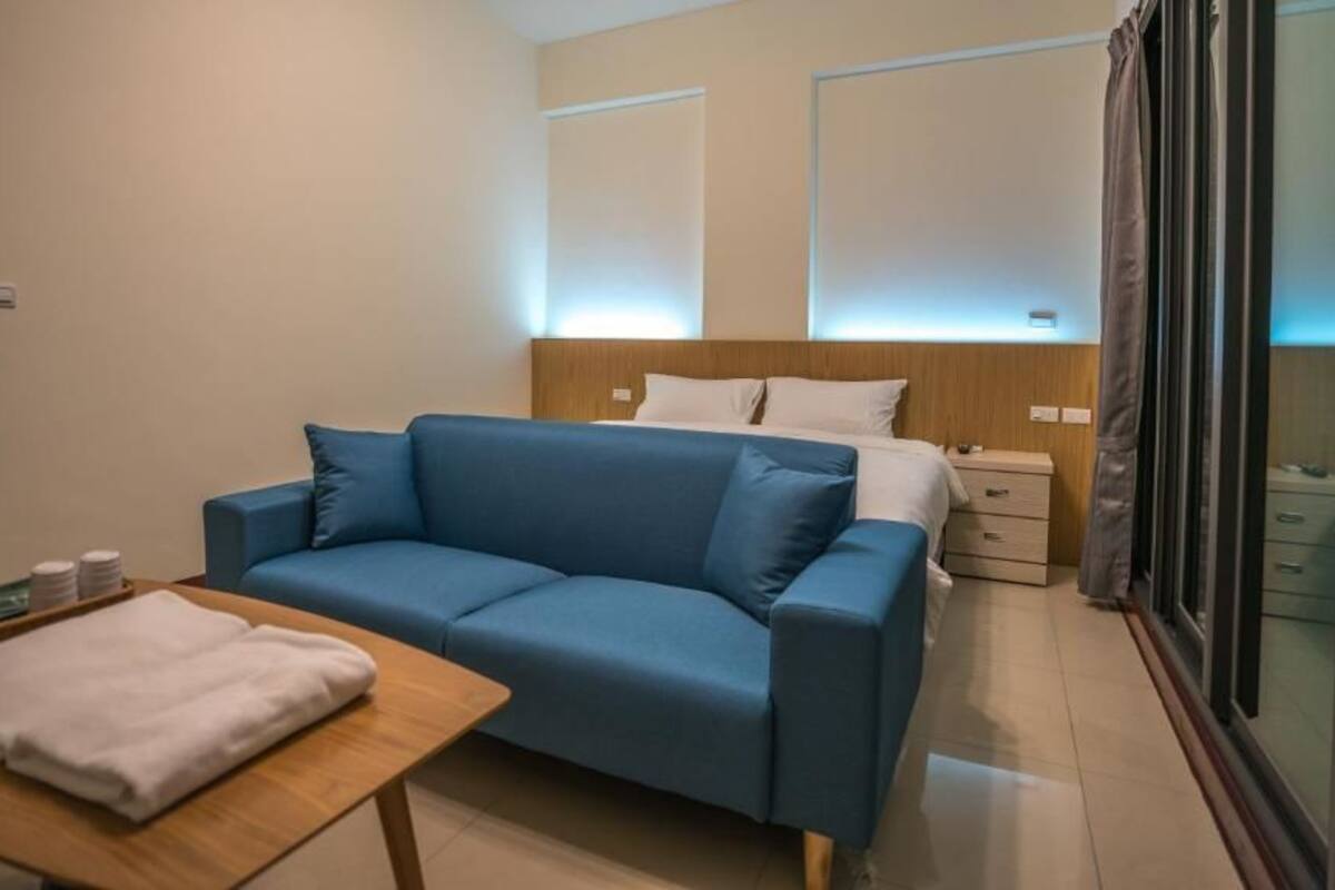 A modern hotel room with a blue sofa, a wooden table with a white towel, a bedside table, and a large window with curtains.