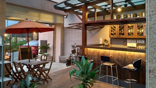An outdoor seating area with wooden tables and chairs, a red umbrella, and a bar area with shelves of bottles. - He-Tian - Cafe & Bar (Puli)