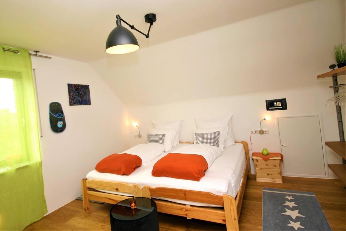 A bedroom with a wooden bed, white bedding, orange pillows, a black lamp, a small table with a green vase, and a star-patterned rug.