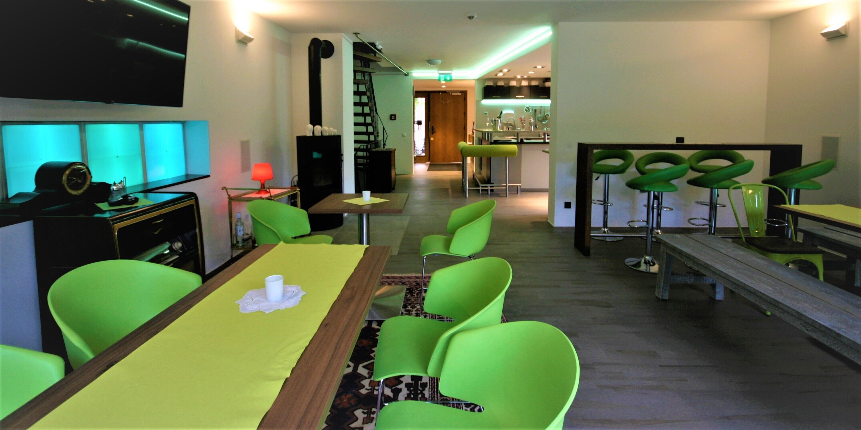 A modern hotel lobby with a long dining table, green chairs, a bar area, and a staircase leading to an upper floor.