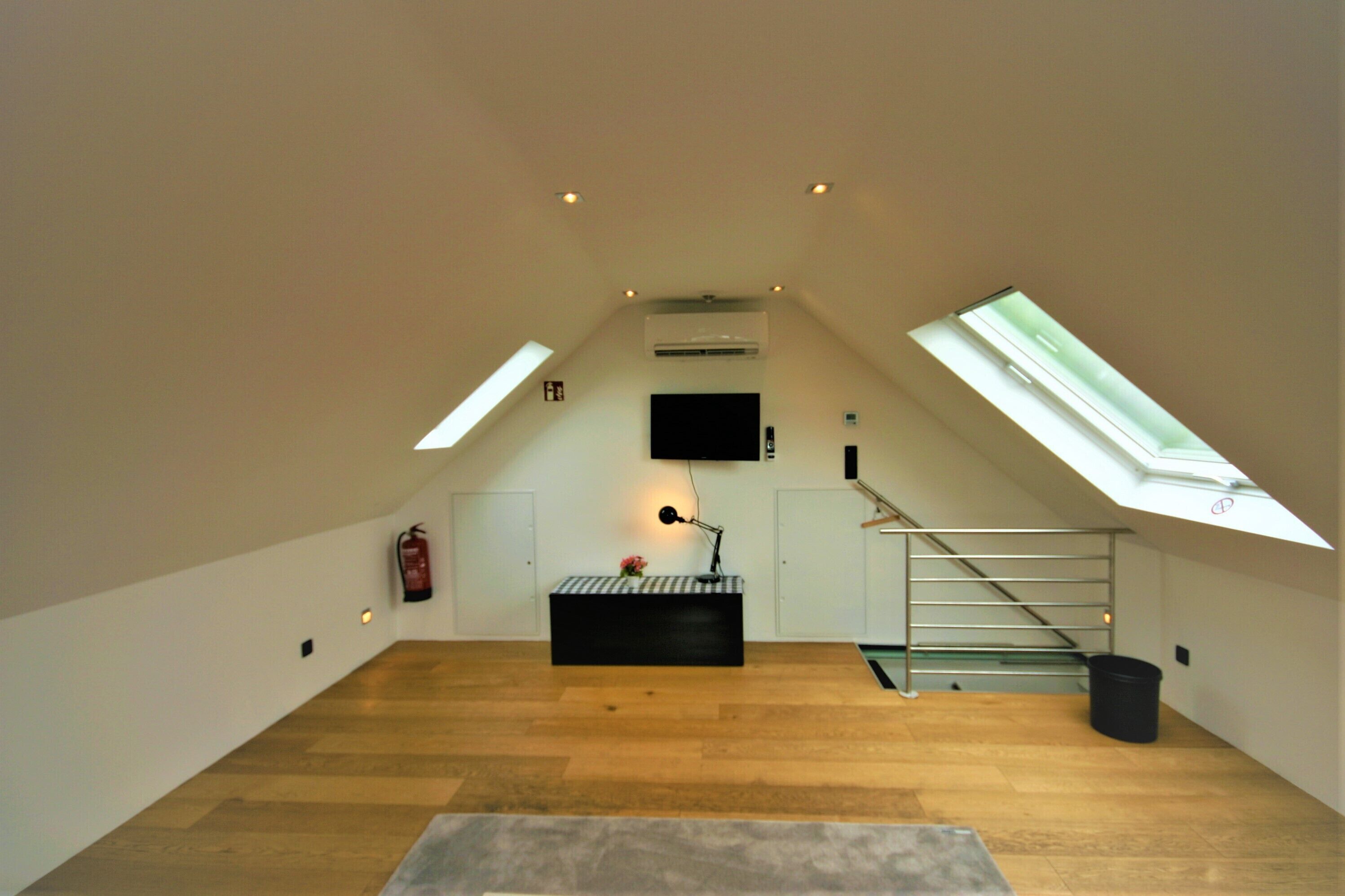 A spacious attic room with wooden flooring, a black bench, a mounted TV, and skylights.