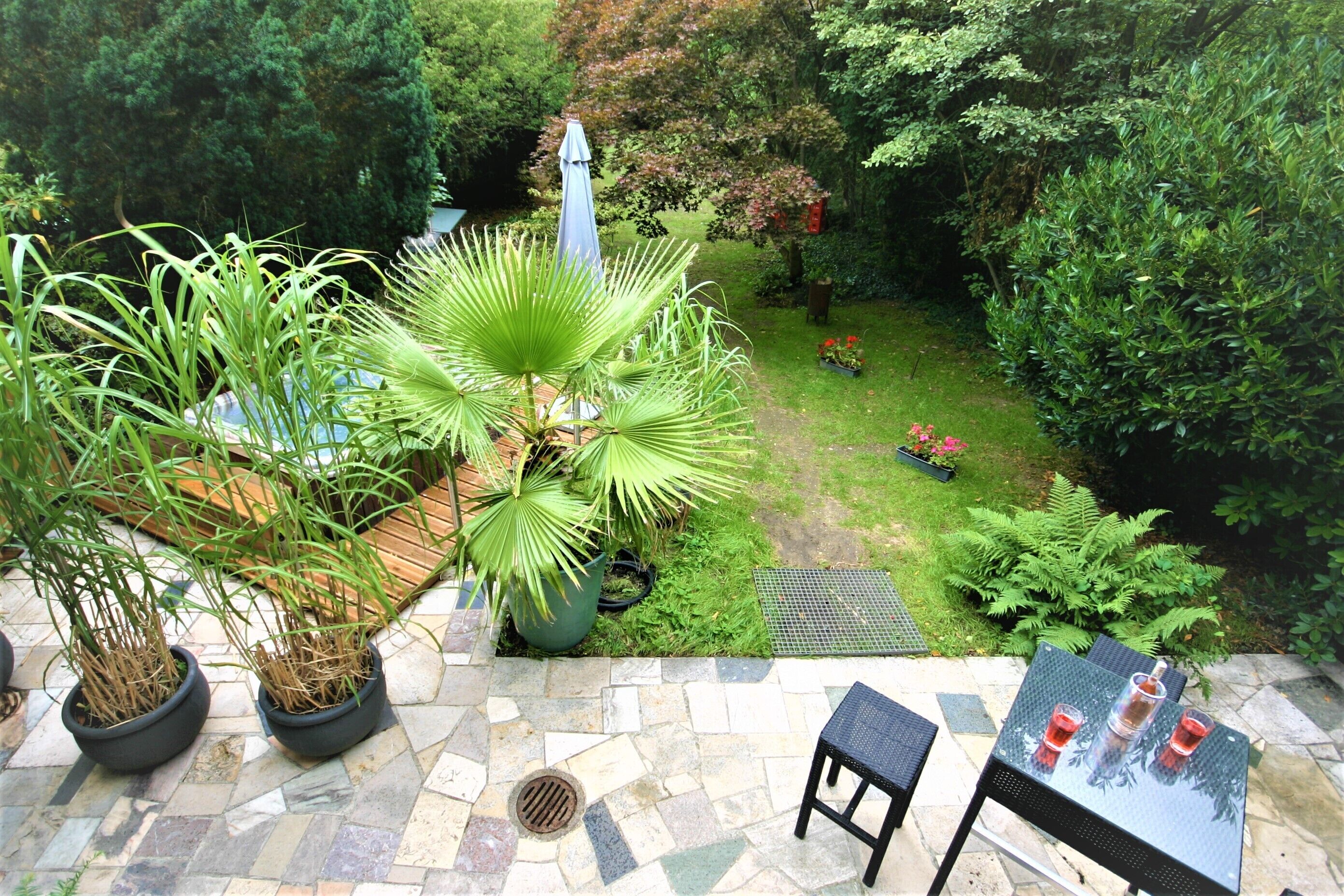 Eine Terrassenfläche mit Steinboden, Blumentöpfen, einem Tisch mit Stühlen und Blick auf einen Garten mit verschiedenen Pflanzen und einer Rasenfläche.