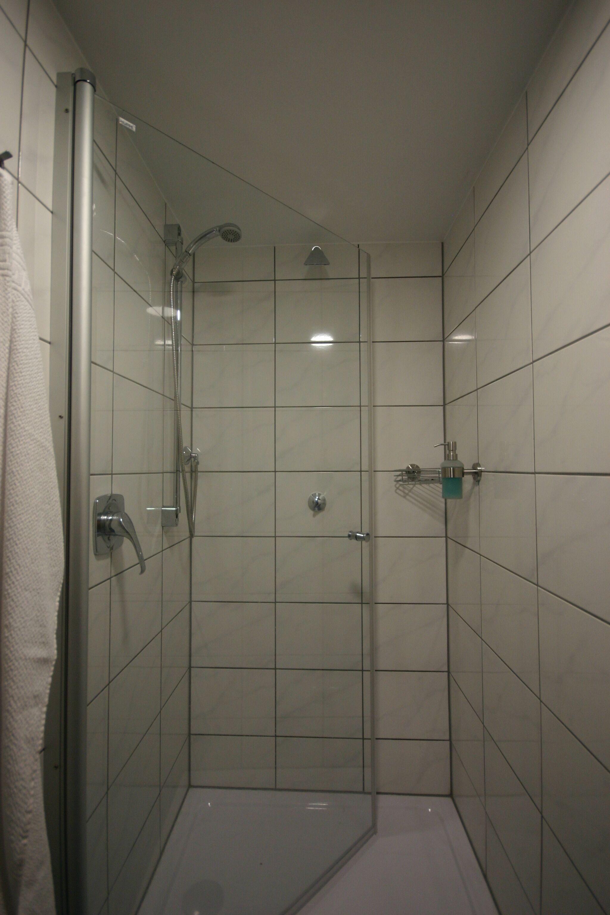 Cabina de ducha con paredes de vidrio, un cabezal de ducha de mano, un grifo de pared y un dispensador de jabón.