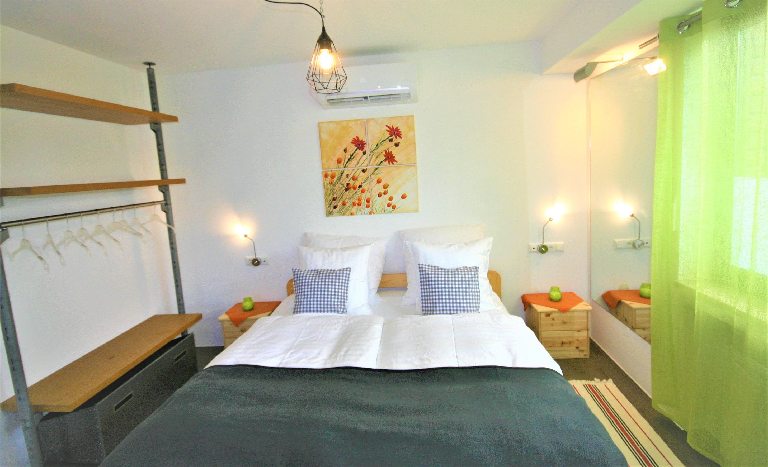 A bedroom with a bed, two bedside tables, a hanging light fixture, a wall-mounted air conditioner, and a green curtain.