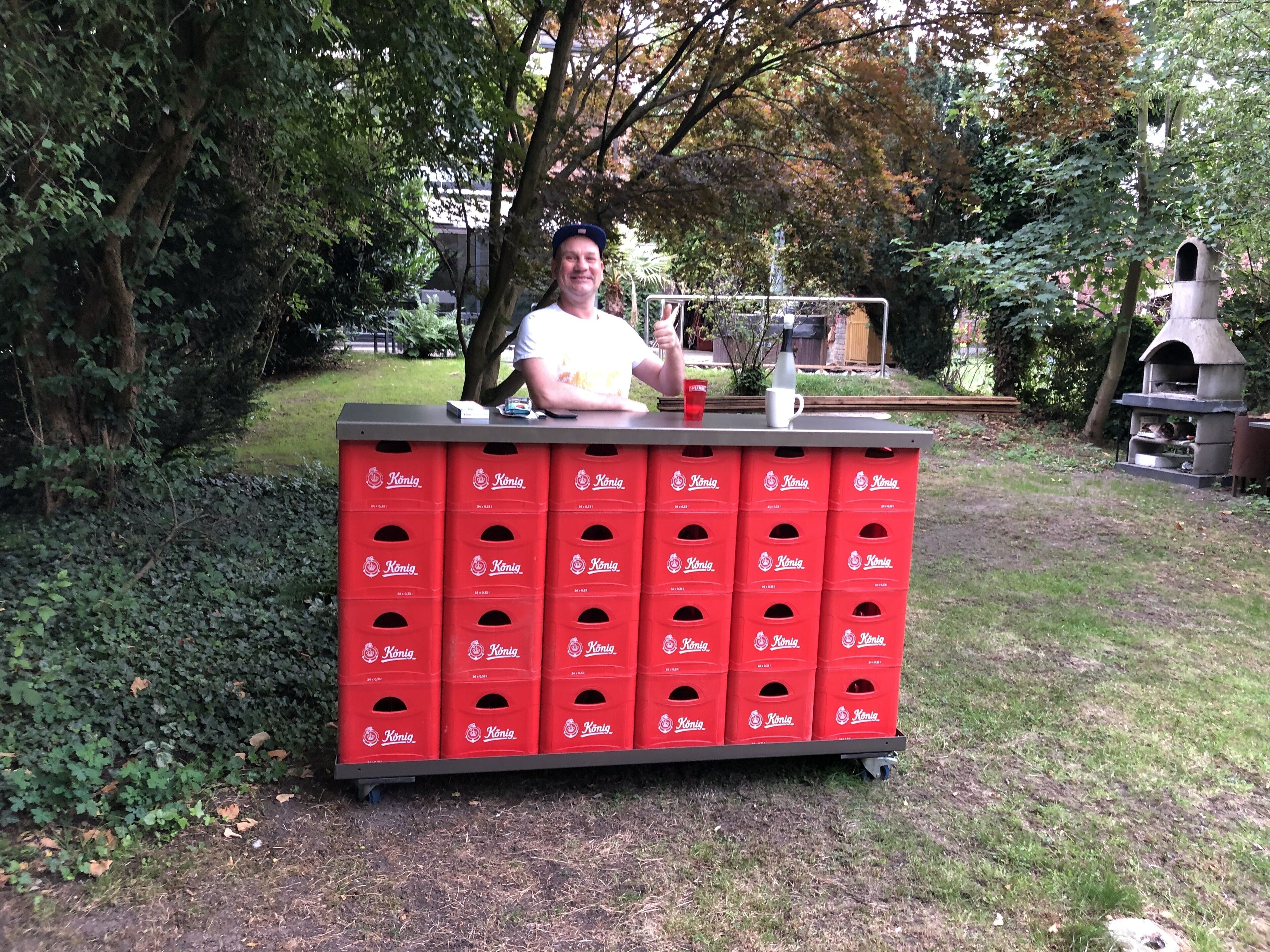 A man standing behind a red beverage dispenser with multiple compartments, set outdoors with trees and a barbecue in the background.