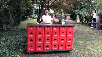 A man standing behind a red beverage dispenser with multiple compartments, set outdoors with trees and a barbecue in the background.