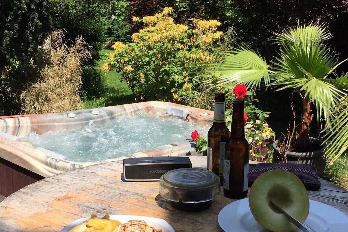 A hot tub surrounded by lush greenery, a wooden table with a bottle, a hat, and a plate with food.