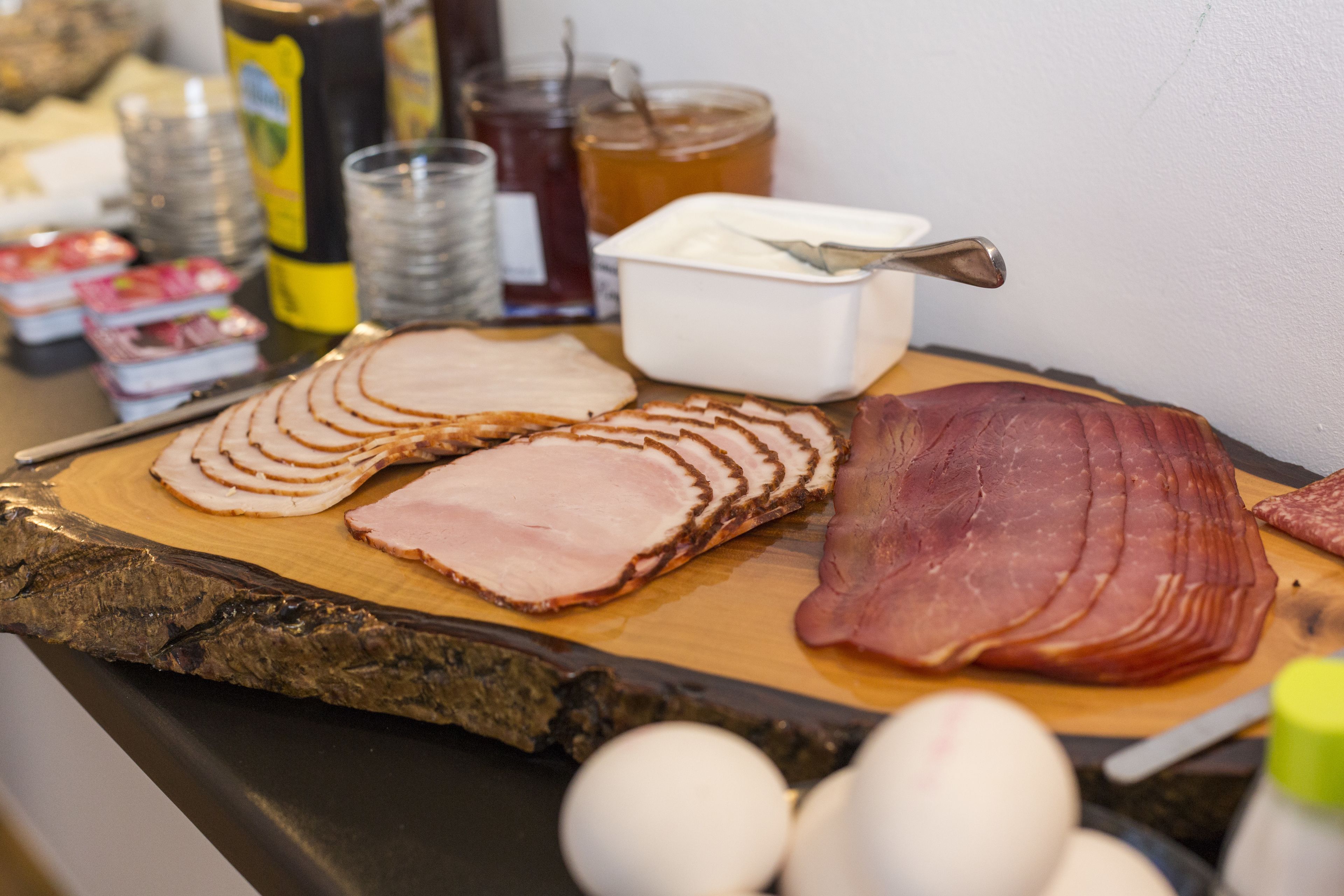 A variety of meats and condiments on a wooden board, including sliced ham and bacon, with eggs and jars of honey and jam visible.