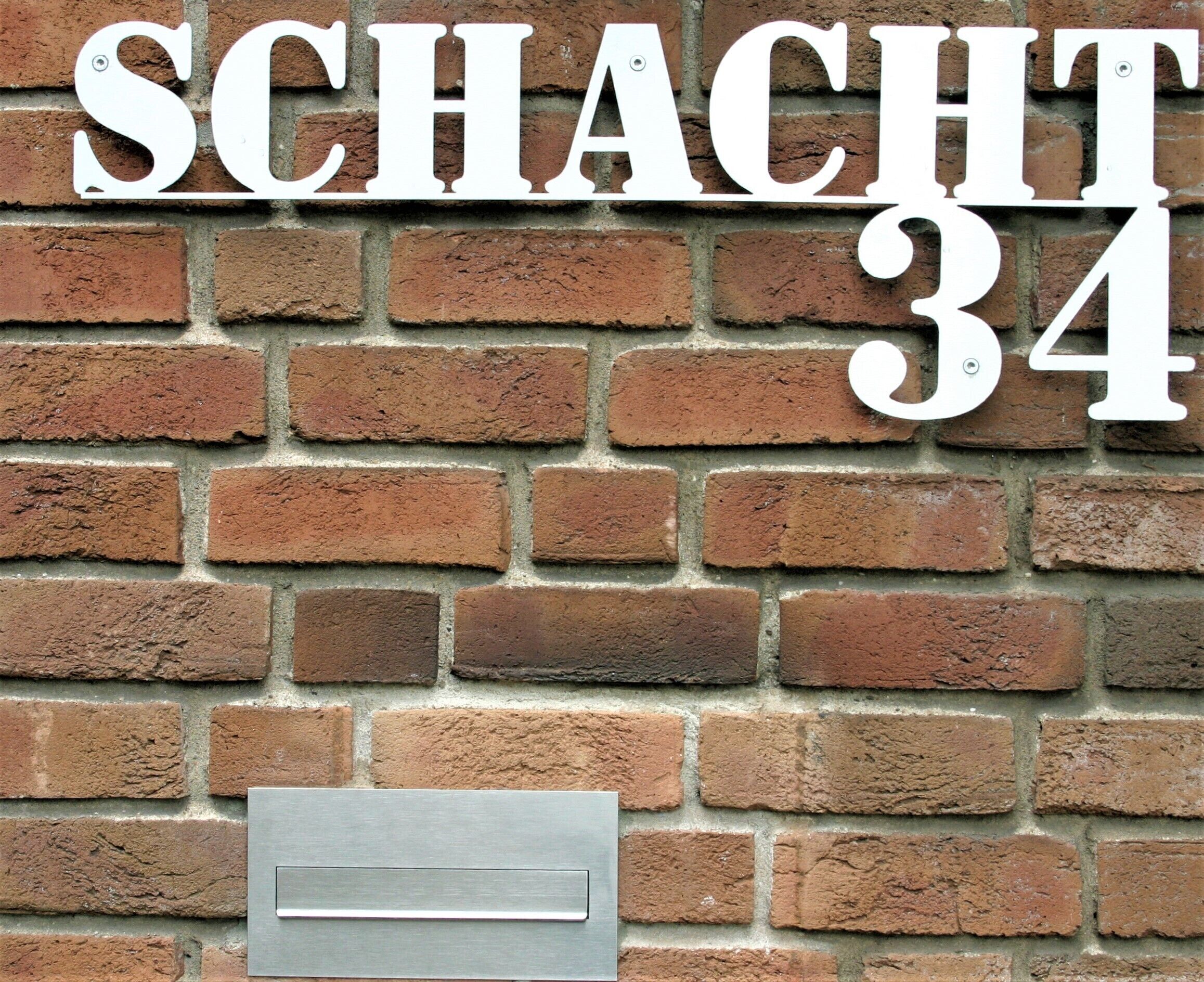 Un mur de briques sur lequel est inscrit le texte « SCHACHT 34 » et où est fixée une boîte aux lettres en métal.