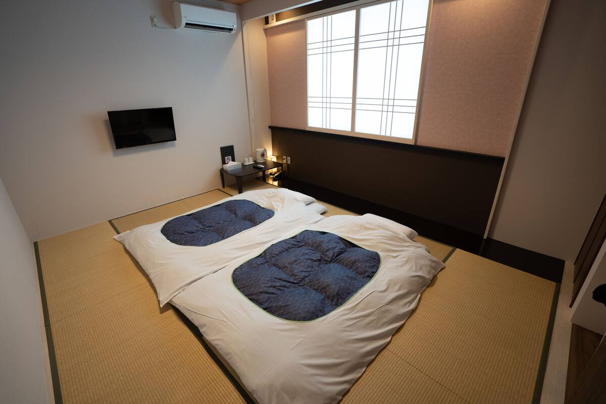 Quarto com piso de tatame, duas camas com roupa de cama azul, uma TV na parede e uma janela com vista.