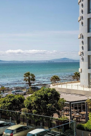 Vista costera con un edificio, una playa y coches aparcados.