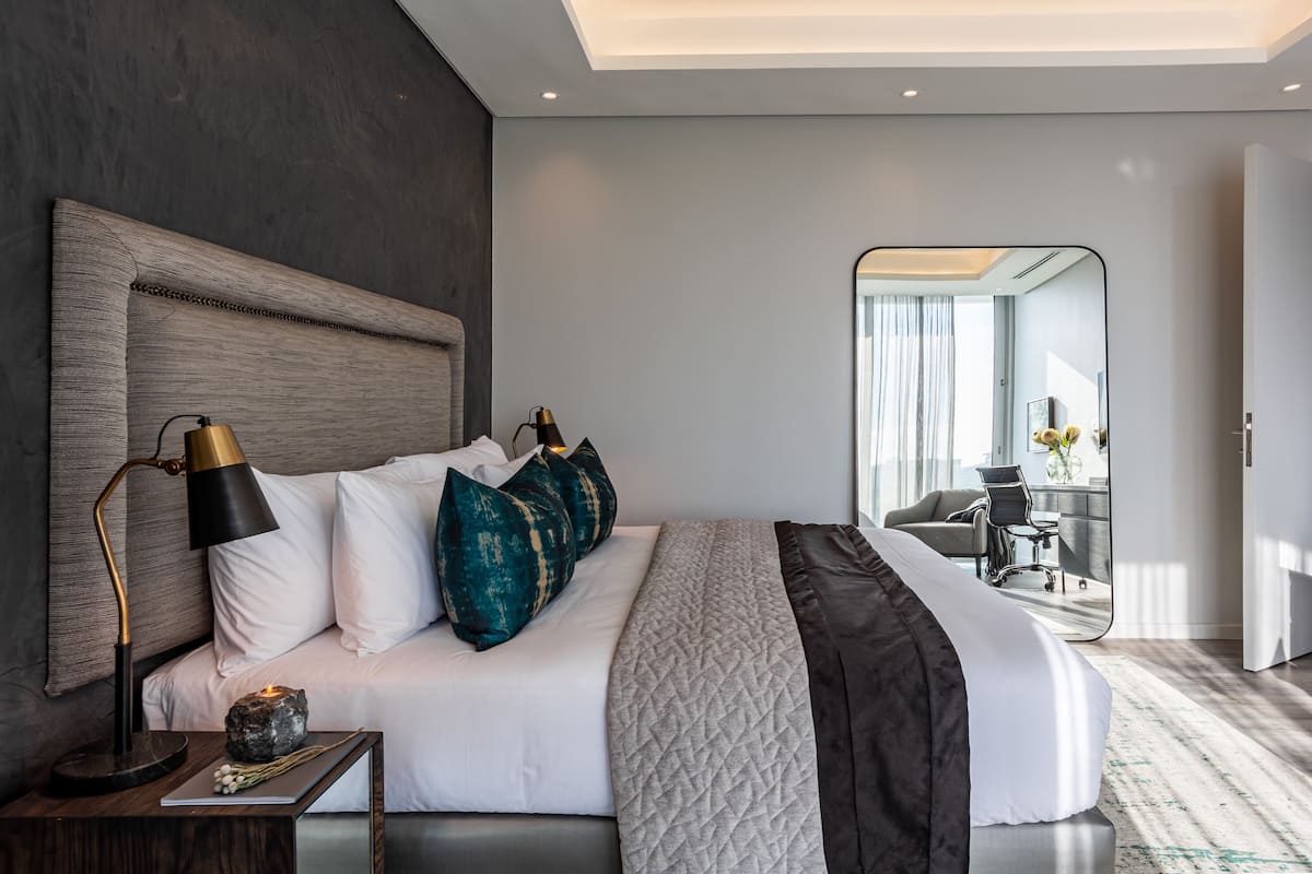 A modern hotel room with a large bed, a bedside table with a lamp, and a view of the adjacent room.