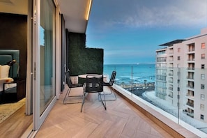 Un balcón con vista al océano, una mesa con sillas y un ventanal que da a una habitación.