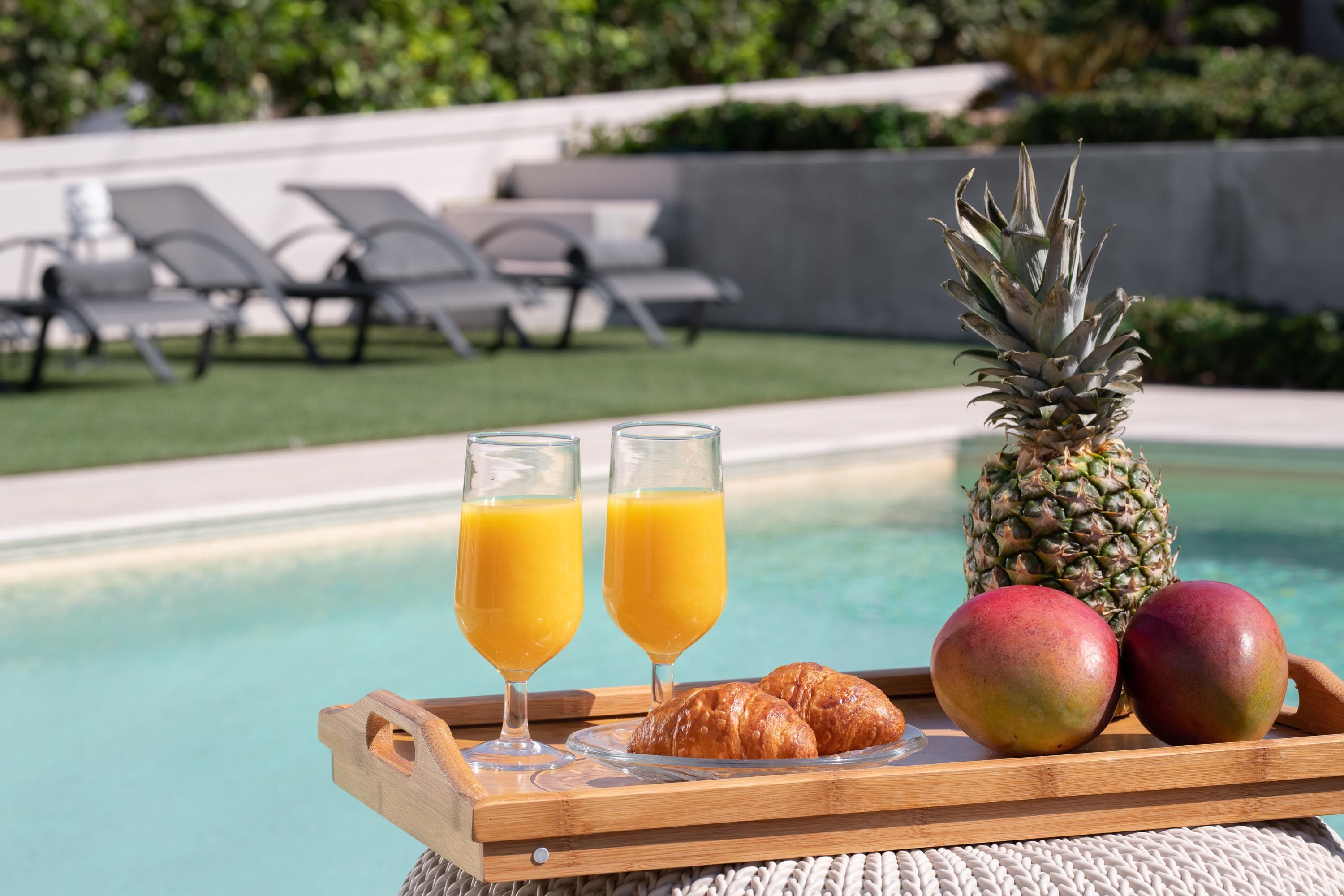 A tray with two glasses of orange juice, a pineapple, a croissant, and two mangoes by a poolside.