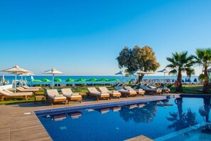 A poolside area with lounge chairs and umbrellas, a clear blue sky, and a view of the ocean. - Cretan Beach Resort - Adults Only (Apokoronas)