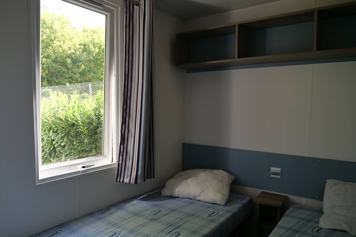 Quarto pequeno com cama de solteiro, janela, cortinas listradas, cabeceira e prateleira sobre a cama.