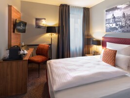 Habitación de hotel con cama, escritorio con cafetera, silla, lámpara y un cuadro de un paisaje urbano.
